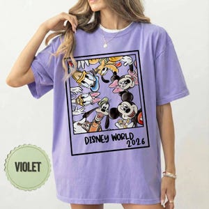 May include: A violet t-shirt with a framed graphic of Disney characters, including Mickey Mouse, Donald Duck, and Goofy. The text "DISNEY WORLD 2026" is printed below. The shirt has short sleeves and a crew neck. The shirt is violet.