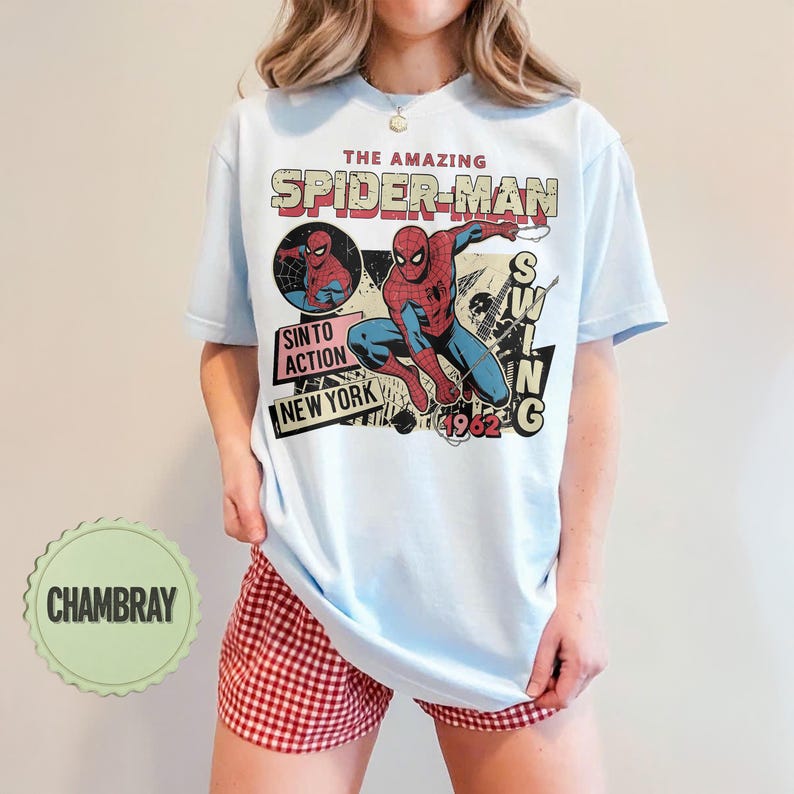 Op de afbeelding: Lichtblauw T-shirt met een vintage afbeelding van Spider-Man. Het ontwerp bevat de tekst "THE AMAZING SPIDER-MAN", "SINTO ACTION NEW YORK" en "1962". Het shirt wordt gecombineerd met een rood-witte geruite short.