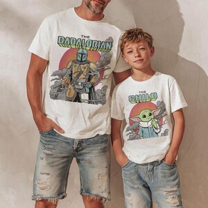 K&ouml;nnte beinhalten: Zwei wei&szlig;e T-Shirts mit kurzen &Auml;rmeln. Das gr&ouml;&szlig;ere Shirt zeigt eine Figur mit Helm und den Text THE DADALORIAN. Das kleinere Shirt zeigt eine gr&uuml;ne Figur mit gro&szlig;en Ohren und den Text THE CHILD. Beide haben einen Hintergrund mit Sonnenuntergang.