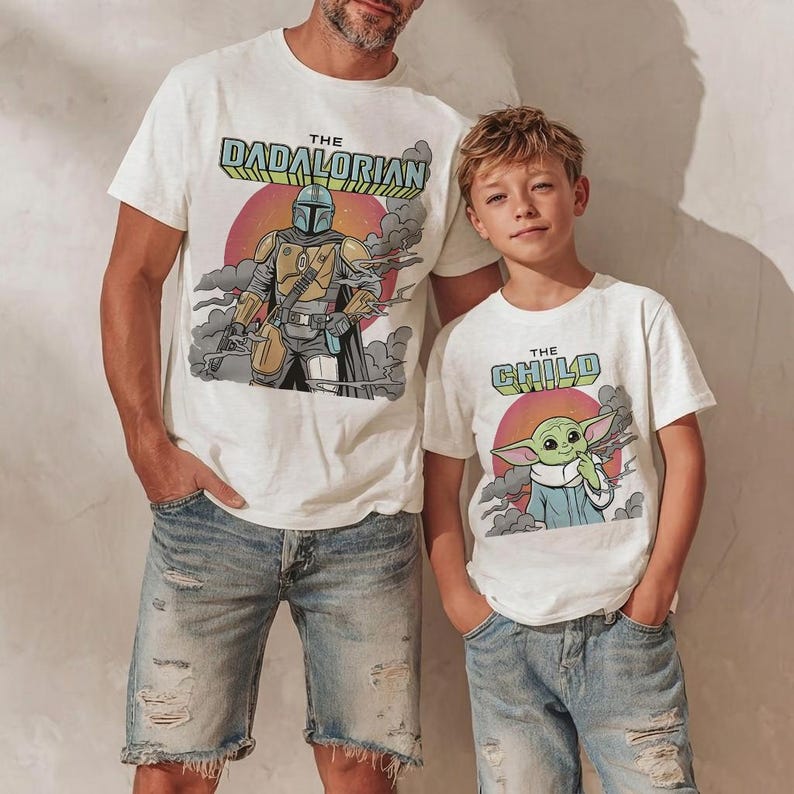 K&ouml;nnte beinhalten: Zwei wei&szlig;e T-Shirts mit kurzen &Auml;rmeln. Das gr&ouml;&szlig;ere Shirt zeigt eine Figur mit Helm und den Text THE DADALORIAN. Das kleinere Shirt zeigt eine gr&uuml;ne Figur mit gro&szlig;en Ohren und den Text THE CHILD. Beide haben einen Hintergrund mit Sonnenuntergang.