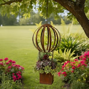 May include: A hanging metal planter with a spherical top and a rectangular base, suspended by a chain from a tree branch. The planter is filled with a variety of succulents in shades of green and purple. The background features a green lawn and flowering plants.