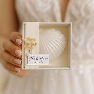 Puede incluir: Una vela blanca en forma de concha en una caja cuadrada, atada con una cinta crema y una ramita de flores secas. Una pequeña etiqueta dice "Eda & Kevin 29.11.2028". La caja es sostenida por una persona.