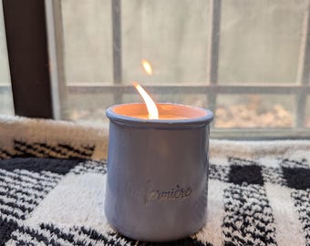 Puede incluir: Una taza de cerámica blanca con asa negra y el texto "But First, Coffee" en negrita negra. La taza se encuentra sobre un fondo borroso, sugiriendo un ambiente acogedor.