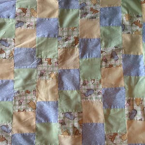 May include: A patchwork quilt with a repeating pattern of squares in pastel shades. The design includes light blue, green, and beige squares, some with polka dots or animal motifs. The quilt's overall aesthetic is soft and inviting.