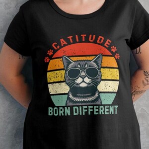 Op de afbeelding: Zwart T-shirt met een kat met zonnebril, tegen een retro zonsondergangontwerp. Het shirt heeft de tekst "CATITUDE" boven de kat en "BORN DIFFERENT" eronder. Het shirt wordt gecombineerd met een witte rok.