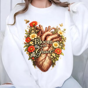 Könnte beinhalten: Weißes Sweatshirt mit einer detaillierten Illustration eines menschlichen Herzens, umgeben von bunten Blumen. Das Design enthält den Text "Bloom with Grace" in eleganter Schrift. Schmetterlinge und Bienen sind ebenfalls im Design enthalten.