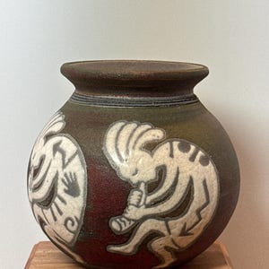 May include: A round, handcrafted ceramic vase with a dark brown and red glaze. The vase features white Kokopelli figures and other Southwestern designs. The vase sits on a wooden block.