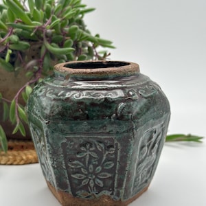 May include: A hexagonal ceramic jar with a dark green, glossy glaze and a brown, unglazed base. The jar features embossed floral patterns on each side, a wide circular opening, and a textured, rustic finish with visible clay edges.