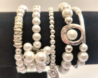 May include: A collection of white pearl bracelets with silver accents. The bracelets feature various designs, including stacked silver rings, large pearls, and a silver circle with a pearl center. A leaf charm hangs from one bracelet.