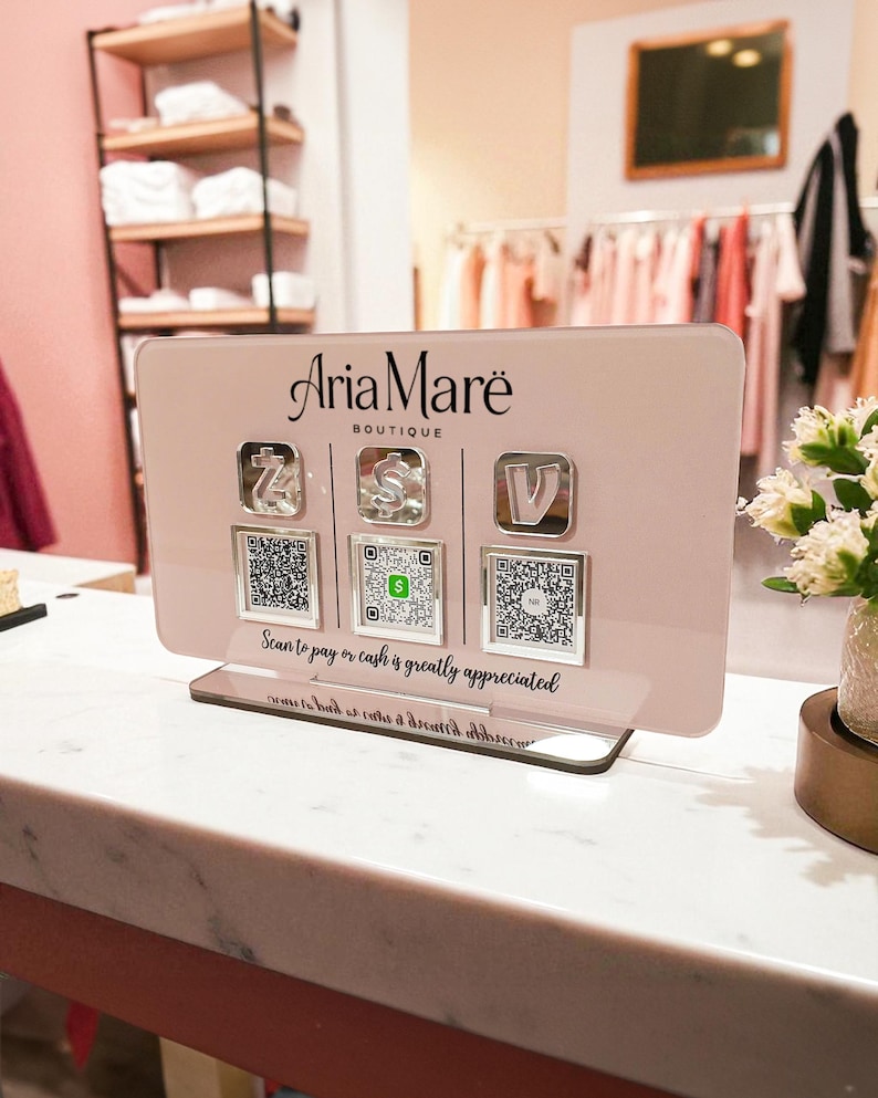 May include: Aria Mar&euml; Boutique payment display. The pink acrylic sign has three payment icons with QR codes below. The text "Scan to pay or cash is greatly appreciated" is printed on the sign.