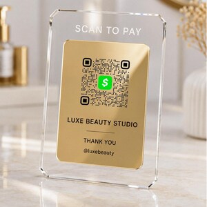 Puede incluir: Un soporte de acrílico transparente para letreros con un inserto rectangular dorado que presenta un código QR negro, un icono de signo de dólar verde y el texto impreso SCAN TO PAY, LUXE BEAUTY STUDIO, THANK YOU y @luxebeauty.