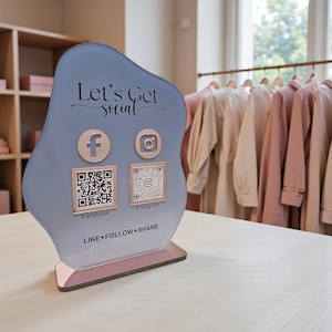 May include: A light blue social media sign with the text "Let's Get Social". It features Facebook and Instagram icons with corresponding QR codes. The base is pink, and the sign says "LIKE &bull; FOLLOW &bull; SHARE".