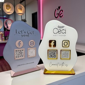 Könnte beinhalten: Zwei dekorative Social-Media-Schilder für ein Nagelstudio. Eines ist hellblau mit dem Text "Let's Get Social" und das andere ist weiß mit dem Text "Ceci nail lounge". Beide haben Facebook- und Instagram-Symbole mit QR-Codes.