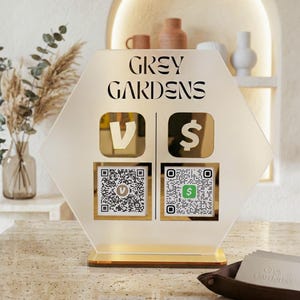 Può includere: Un cartello esagonale con la scritta "GREY GARDENS" e icone di pagamento. Il cartello ha due codici QR e una base dorata. Lo sfondo include oggetti decorativi e un vassoio con biglietti da visita.