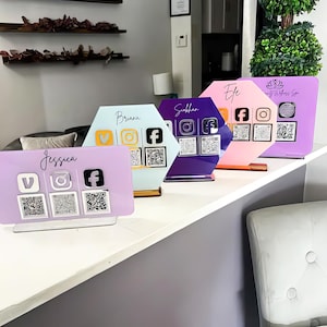 Puede incluir: Una fila de coloridas pantallas acrílicas de redes sociales. Cada pantalla presenta un nombre diferente, iconos de redes sociales y códigos QR. Los colores incluyen lavanda, azul claro, morado, rosa y una última pantalla lavanda. Las pantallas están sobre una superficie blanca.