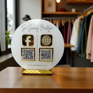 May include: A white and gold sign for So Savvy Boutique, featuring the Facebook and website logos, along with QR codes. The sign is on a wooden table, with clothing racks visible in the background. The website address, Sosavvyboutique.com, is printed at the bottom.
