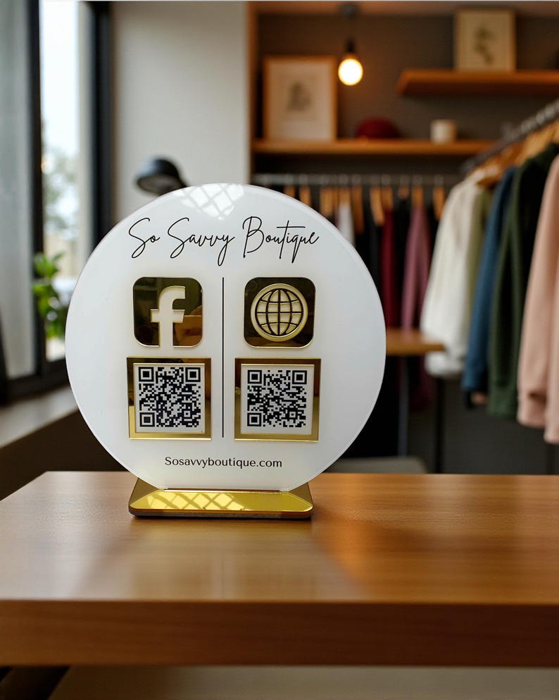 May include: A white and gold sign for So Savvy Boutique, featuring the Facebook and website logos, along with QR codes. The sign is on a wooden table, with clothing racks visible in the background. The website address, Sosavvyboutique.com, is printed at the bottom.
