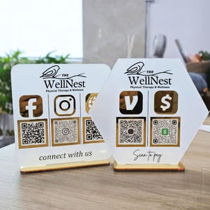 May include: Two acrylic signs with gold trim. One is rectangular, the other is hexagonal. Both feature the text "The WellNest Physical Therapy & Wellness" and social media icons with QR codes. One sign says "connect with us" and the other says "Scan to pay".