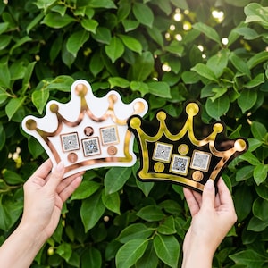 May include: Two crown-shaped social media signs, one white with rose gold accents and the other black with gold accents. Each crown has QR codes and social media icons. The background is a lush green hedge.