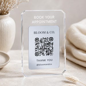 Puede incluir: Un letrero rectangular de acrílico transparente con bordes biselados que presenta el texto blanco BOOK YOUR APPOINTMENT en la parte superior. El centro muestra una tarjeta blanca con el texto negro BLOOM & CO., un código QR, THANK YOU y @bloomandco.