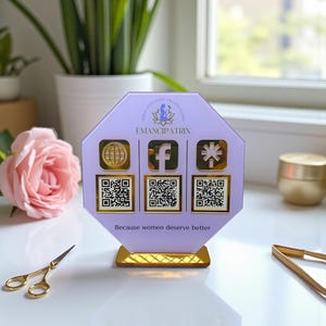 May include: A lavender acrylic display with gold accents. The display features the text "EMANCIPATRIX" and social media icons with QR codes. The phrase "Because women deserve better" is printed at the bottom.