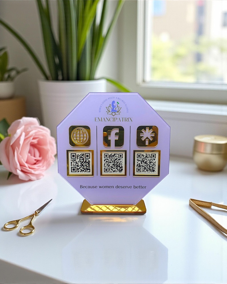 May include: A lavender acrylic display with gold accents. The display features the text "EMANCIPATRIX" and social media icons with QR codes. The phrase "Because women deserve better" is printed at the bottom.