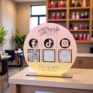以下が含まれることがあります： Bre's Beauty Loungeの丸い照明付きサイン。Facebook、TikTok、Instagramのソーシャルメディアアイコンと各QRコードが表示されています。サインはピンクと黄色のグラデーションの背景で、「Thank you for supporting my small business」というテキストがあります。
