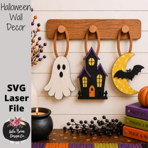 May include: Three wooden Halloween wall decorations hanging from a horizontal wooden rack. The ornaments include a white ghost, a black haunted house with yellow windows, and a yellow crescent moon with a black bat. Text reads Halloween Wall Decor.