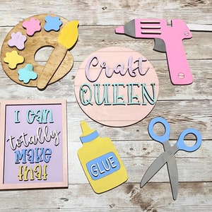 May include: A set of wooden craft signs with a pink, blue, and yellow color scheme. The signs feature phrases like "Craft Queen", "I can totally make that", and "Glue". There is also a paint palette with a paintbrush, a glue bottle, and a pair of scissors.