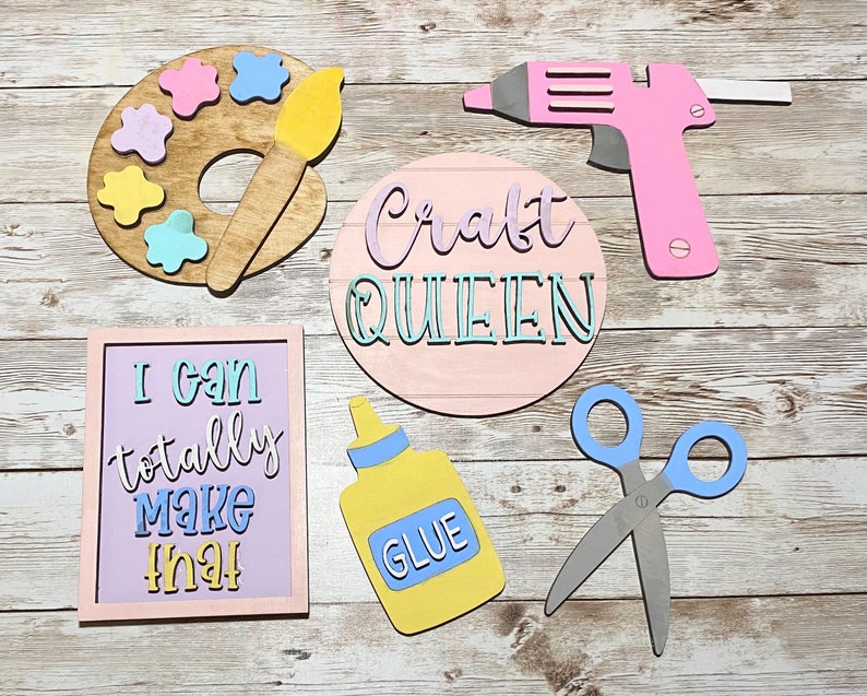 May include: A set of wooden craft signs with a pink, blue, and yellow color scheme. The signs feature phrases like "Craft Queen", "I can totally make that", and "Glue". There is also a paint palette with a paintbrush, a glue bottle, and a pair of scissors.