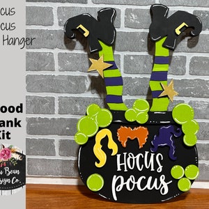 May include: A wooden door hanger kit featuring a black cauldron with green bubbles, witch legs with striped stockings and black boots, and the words "Hocus Pocus".