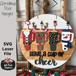 Pode incluir: Cabide de porta de Natal em madeira com quatro desenhos de canecas de caf&eacute; festivas: Papai Noel, rena, uma caneca com luzes de Natal e um boneco de neve. O texto "Have a cup of cheer" est&aacute; abaixo. Arquivo a laser.