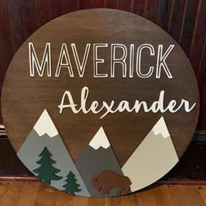 K&ouml;nnte beinhalten: Ein rundes Holzschild mit den Namen "Maverick Alexander" in wei&szlig;en Buchstaben. Das Schild zeigt eine Berglandschaft mit gr&uuml;nen B&auml;umen und einem braunen Bison.