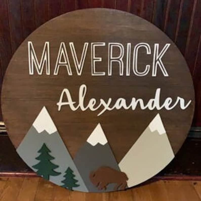 K&ouml;nnte beinhalten: Ein rundes Holzschild mit den Namen "Maverick Alexander" in wei&szlig;en Buchstaben. Das Schild zeigt eine Berglandschaft mit gr&uuml;nen B&auml;umen und einem braunen Bison.