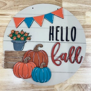 Puede incluir: Un letrero redondo de madera con las palabras "Hello Fall" en letras negras y rojas. El letrero est&aacute; decorado con una hilera de banderas naranja y turquesa, una planta en maceta con flores naranjas y tres calabazas: una naranja, una naranja con tallos verdes y una turquesa.