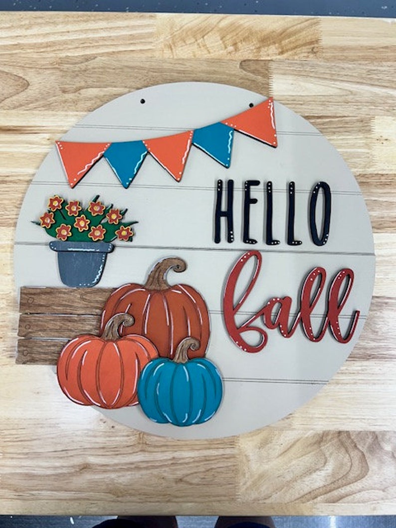 Puede incluir: Un letrero redondo de madera con las palabras "Hello Fall" en letras negras y rojas. El letrero est&aacute; decorado con una hilera de banderas naranja y turquesa, una planta en maceta con flores naranjas y tres calabazas: una naranja, una naranja con tallos verdes y una turquesa.