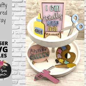 May include: A white tiered tray with various craft-themed decorations, including a pink sign that says "I can totally make that", a pink sign that says "Craft Queen", a paint palette, a glue bottle, scissors, and a hot glue gun.