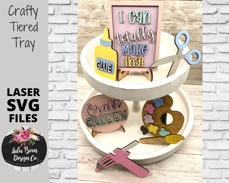 May include: A white tiered tray with various craft-themed decorations, including a pink sign that says "I can totally make that", a pink sign that says "Craft Queen", a paint palette, a glue bottle, scissors, and a hot glue gun.