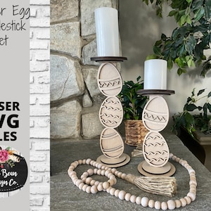 Może przedstawiać: Zestaw dwóch drewnianych świeczników w kształcie jajek wielkanocnych. Każdy świecznik składa się z trzech ułożonych na sobie jajek z różnymi wzorami. Jajka są pomalowane na biało i mają naturalne wykończenie drewna. Świeczniki są ustawione na stole z drewnianym naszyjnikiem z koralików.