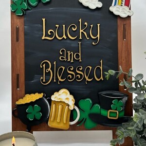 May include: A wooden chalkboard sign with a black background and the words "Lucky and Blessed" in gold lettering. The sign is decorated with a rainbow, shamrocks, a pot of gold, a beer mug, and a leprechaun's hat.