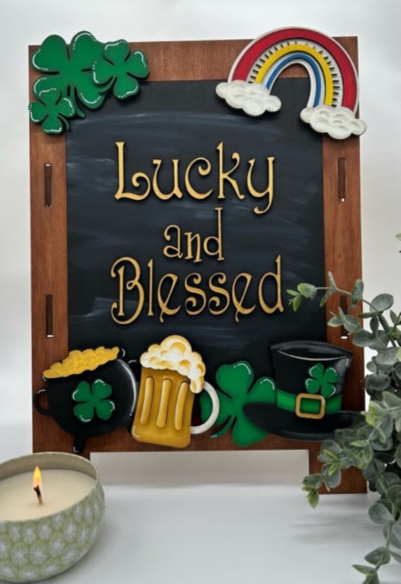 May include: A wooden chalkboard sign with a black background and the words "Lucky and Blessed" in gold lettering. The sign is decorated with a rainbow, shamrocks, a pot of gold, a beer mug, and a leprechaun's hat.