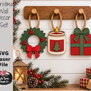 May include: A Christmas wall decor set featuring three wooden hanging ornaments: a green wreath with a red bow, a candle in a jar with a tree graphic, and a red gift box with a green bow, all suspended from a horizontal wooden rack with three pegs.