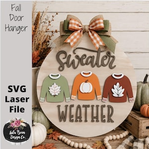 May include: A round wooden door hanger featuring the text Sweater Weather in dark brown script and block letters. It displays three wooden sweater cutouts in sage green, orange, and rust, decorated with a white leaf and pumpkin, topped with a plaid bow.