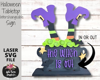 May include: A Halloween tabletop sign with a witch's legs sticking out of a cauldron. The sign says "The Witch Is Out" and has a black and white bat design. The sign is interchangeable and can be used to say "In" or "Out".