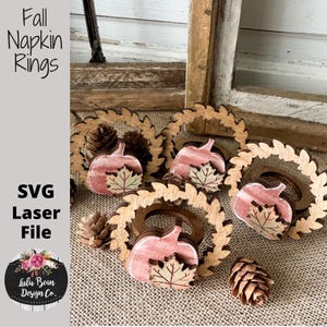 May include: Wooden fall-themed napkin rings with pumpkin and leaf designs. The rings are arranged with pinecones on a burlap surface. The image includes the text "Fall Napkin Rings" and "SVG Laser File".