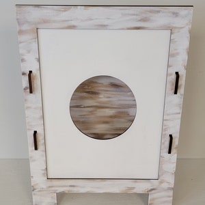 Op de afbeelding: Wit houten kastje met een ronde uitsparing en een verweerde verfbehandeling. Het kastje heeft vier kleine houten pootjes.