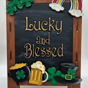 May include: A wooden chalkboard easel with a black chalkboard surface. The chalkboard has the words "Lucky and Blessed" written in gold paint. The easel is decorated with a rainbow, shamrocks, a pot of gold, a beer mug, and a leprechaun hat.
