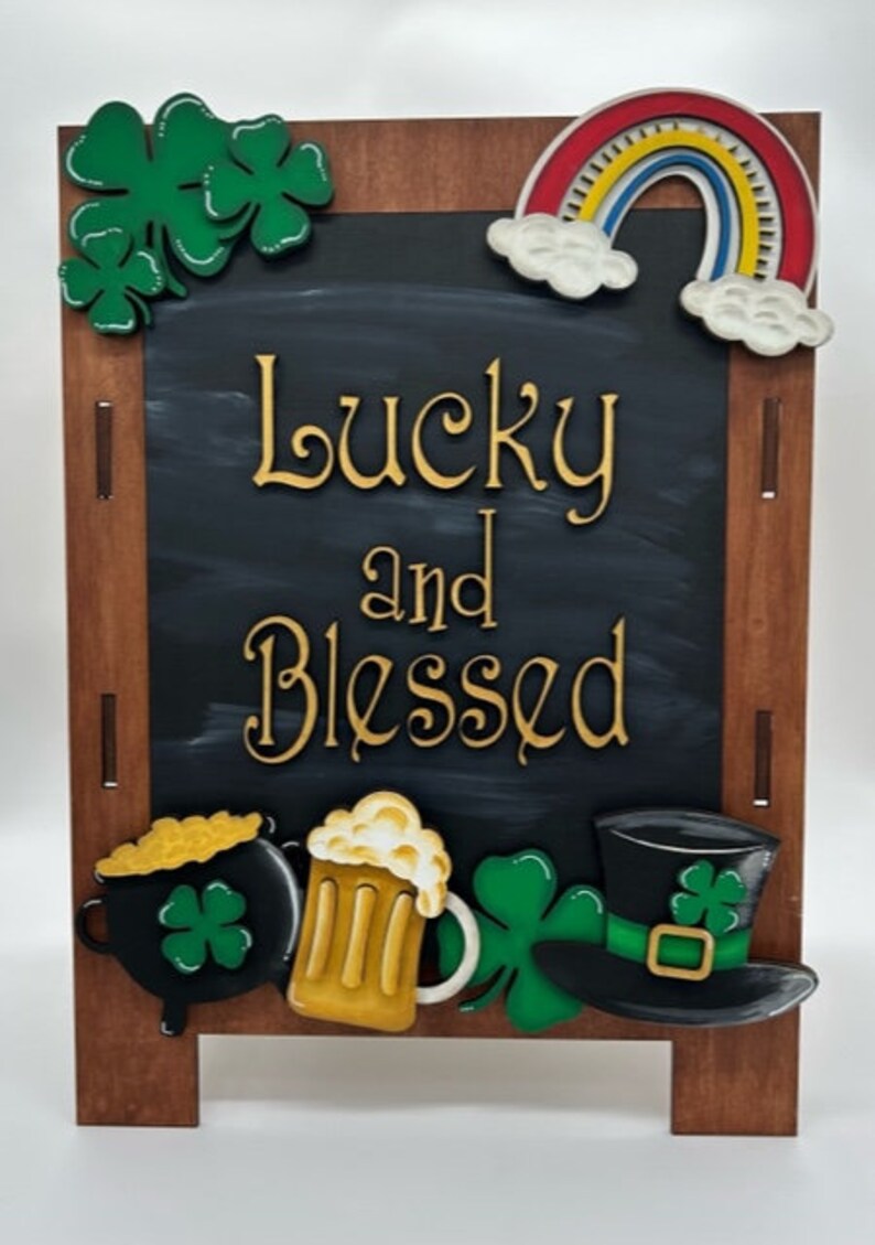May include: A wooden chalkboard easel with a black chalkboard surface. The chalkboard has the words "Lucky and Blessed" written in gold paint. The easel is decorated with a rainbow, shamrocks, a pot of gold, a beer mug, and a leprechaun hat.