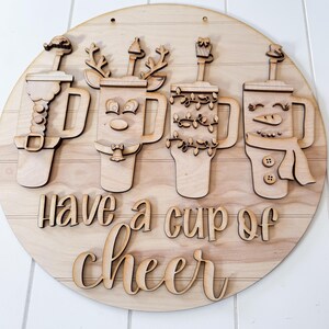 Pode incluir: Um letreiro redondo de madeira com a frase "Have a cup of cheer". O letreiro apresenta quatro canecas com decora&ccedil;&otilde;es tem&aacute;ticas de feriados, incluindo um boneco de neve, uma rena e luzes de Natal. O letreiro &eacute; feito de madeira clara.