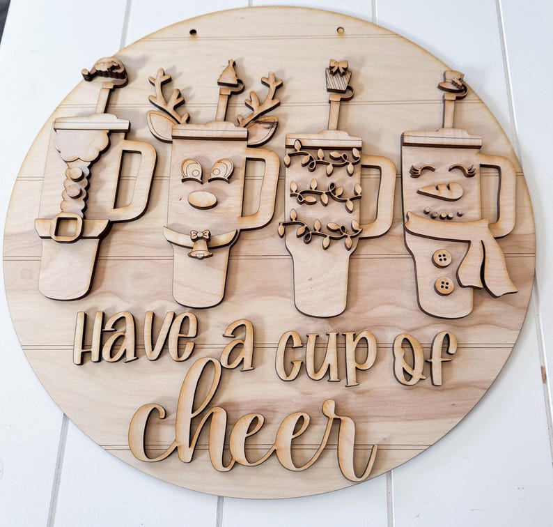 Pode incluir: Um letreiro redondo de madeira com a frase "Have a cup of cheer". O letreiro apresenta quatro canecas com decora&ccedil;&otilde;es tem&aacute;ticas de feriados, incluindo um boneco de neve, uma rena e luzes de Natal. O letreiro &eacute; feito de madeira clara.